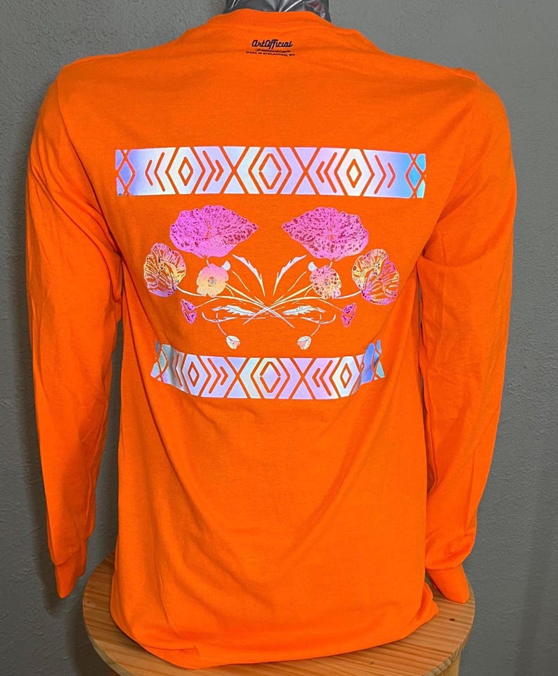 May include: Orange long-sleeve t-shirt with a reflective design on the back. The design features a floral pattern with pink and purple flowers and a geometric pattern with white and blue accents. The text "ArtOfficial" is printed on the back of the shirt.