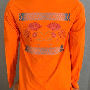 May include: Orange long-sleeve t-shirt with a geometric design and floral pattern on the back. The design features a gray and white geometric pattern with a floral pattern in shades of red, pink, and orange. The shirt has the text "Old/Faded" on the back.