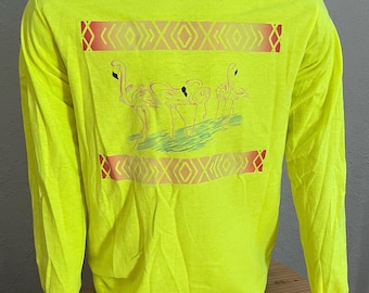 Puede incluir: Una camiseta de manga larga de color amarillo neón con un gráfico de flamencos rosas de pie en el agua. El gráfico está rodeado por un patrón geométrico rojo y naranja.