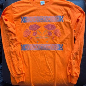 May include: Orange long-sleeve t-shirt with a floral design and a geometric pattern. The design features pink and red flowers with green stems and leaves. The geometric pattern is in shades of blue and gray.