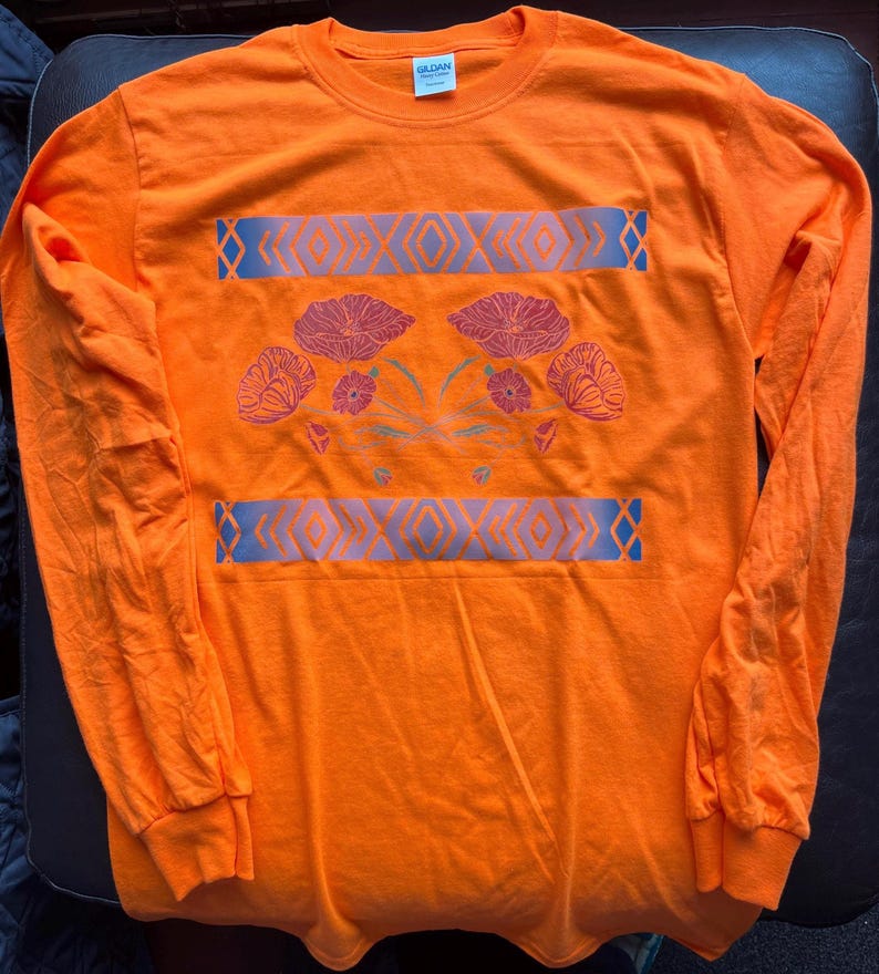 May include: Orange long-sleeve t-shirt with a floral design and a geometric pattern. The design features pink and red flowers with green stems and leaves. The geometric pattern is in shades of blue and gray.