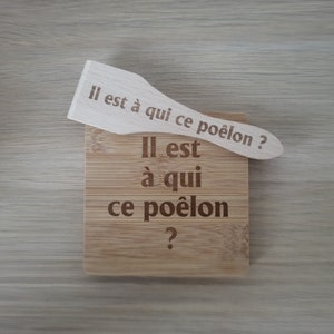 Könnte beinhalten: Ein Holzspatel mit dem Text "Il est à qui ce poêlon?" eingraviert. Der Spatel liegt auf einem Holzbrett mit dem gleichen Text eingraviert.
