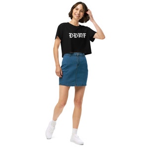 May include: A woman wearing a black cropped t-shirt with the text "DYWMF" in white gothic lettering. She is also wearing a blue denim skirt and white sneakers.