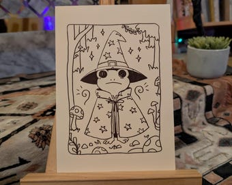 May include: A black and white line drawing of a frog wearing a wizard hat and robe with stars. The frog is standing in a forest setting with mushrooms and stars.