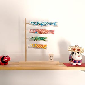 May include: A wooden shelf with a white edge displays a set of colourful fish-shaped windsocks, a red figurine, and a white figurine wearing a straw hat. The windsocks are hanging from a wooden stand. The shelf is decorated with a small wooden block with a symbol.
