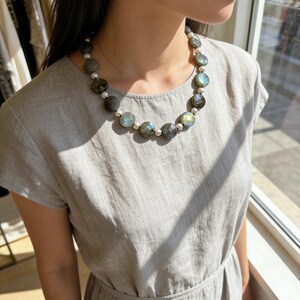 May include: A necklace featuring large, round, gray-green labradorite stones interspersed with small, white pearls. The necklace is worn against a light gray dress. The labradorite stones have iridescent blue and green flashes.