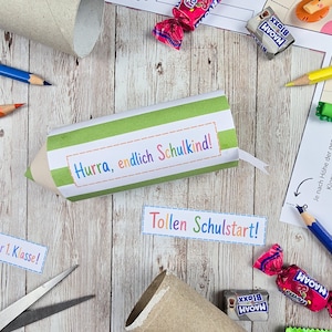 Op de afbeelding: Een cilindervormig papieren knutselwerk in de vorm van een potlood met groene en witte strepen, voorzien van de tekst Hurra, endlich Schulkind! in kleurrijke letters. Daarnaast liggen papieren uitknipsels met de tekst Tollen Schulstart! en 1. Klasse!, snoepjes en potloden.