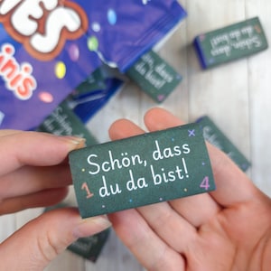 Könnte beinhalten: Eine rechteckige, dunkelgrüne Bonbonverpackung mit dem weißen Text "Schön, dass du da bist!" und kleinen bunten Akzenten, darunter eine pfirsichfarbene 1 und eine rosa 4, umgeben von weiteren identischen Verpackungen und einer blauen Tüte.