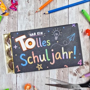 May include: A rectangular black box containing round chocolates, featuring the colorful text "HAB EIN Tolles Schuljahr!" with illustrations of a lightbulb, a paper airplane, stars, and numbers, surrounded by pencils, scissors, and candy.