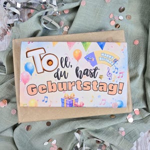 May include: A rectangular birthday card featuring the German text "Toll, du hast GEBURTSTAG!" in large, colorful letters. The design includes illustrations of balloons, a gift box, musical notes, and a fan-shaped "Happy Birthday" banner.
