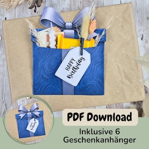 Può includere: Una busta blu a forma di regalo con un grande fiocco viola, contenente una candela bianca, un oggetto rettangolare giallo ed erbe essiccate. Un'etichetta bianca fissata con una clip nera riporta il testo nero "HAPPY BIRTHDAY".
