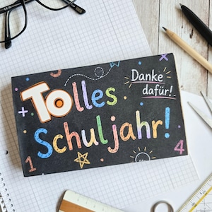Könnte beinhalten: Ein rechteckiges schwarzes Schild mit buntem, handgeschriebenem Text, der Tolles Schuljahr! und Danke dafür! in verschiedenen leuchtenden Farben zeigt. Das Design enthält kleine handgezeichnete Sterne, Zahlen und Kritzeleien.