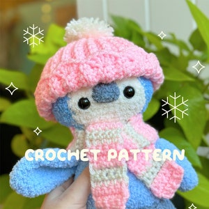 A cute blue crochet penguin wearing a pink crochet hat with a white pom pom and a pink and white striped crochet scarf.  The photo has text on it which says "crochet pattern by @snailmail.studios"