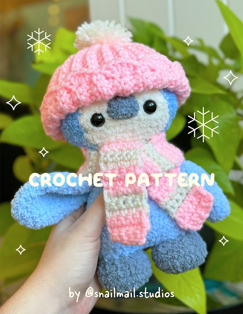 A cute blue crochet penguin wearing a pink crochet hat with a white pom pom and a pink and white striped crochet scarf.  The photo has text on it which says "crochet pattern by @snailmail.studios"