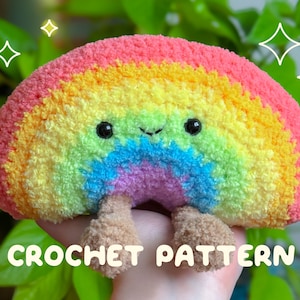 Puede incluir: Un juguete de peluche arcoíris de crochet con una cara sonriente y dos pequeñas piernas. El arcoíris está hecho de hilo de colores en tonos de rojo, naranja, amarillo, verde, azul, añil y violeta. El juguete está siendo sostenido en una mano con el texto "CROCHET PATTERN" debajo.