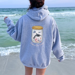May include: A light gray hoodie featuring a watercolor illustration of marine life inside a jar. The jar contains a whale, turtle, and other sea creatures. The words "Preserve Our Oceans" are printed below the jar in a cursive font.