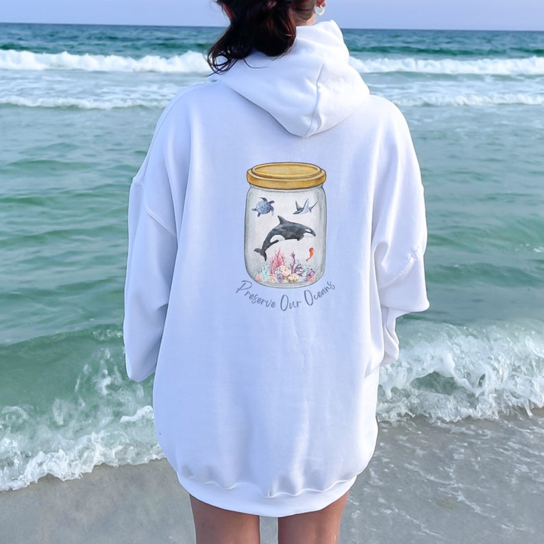 May include: White hoodie with a graphic of marine life inside a jar, including a turtle, orca, and coral. The text "Preserve Our Oceans" is printed below the jar. The hoodie is worn by a person at the beach.