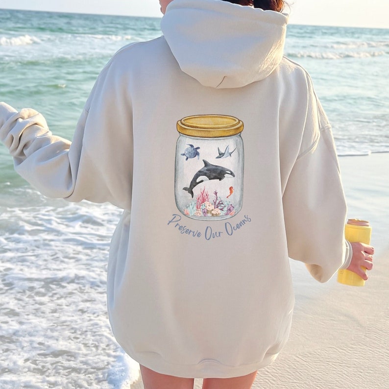 May include: Beige hoodie with a watercolor illustration of marine life inside a jar. The jar contains a whale, turtle, and other sea creatures. The text "Preserve Our Oceans" is below the jar.