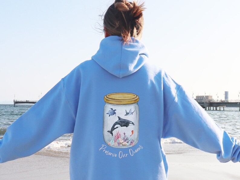May include: Light blue hoodie with a watercolor illustration of ocean life inside a jar. The jar contains a turtle, orca, stingray, and coral. The text "Preserve Our Oceans" is below the jar.
