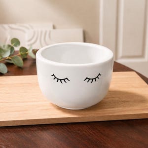 May include: A white ceramic bowl with black eyelash designs. The bowl is round with a smooth surface and is sitting on a wooden tray. The background includes a wooden surface and a white wall.