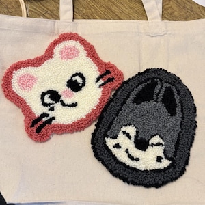 Op de afbeelding: Een beige canvas tas met twee punch needle patches. De ene patch is een wit kattengezicht met roze oren en een rode rand. De tweede patch is een grijs wolfsgezicht met een zwarte rand. Beide patches hebben een getextureerde, lussenstructuur.
