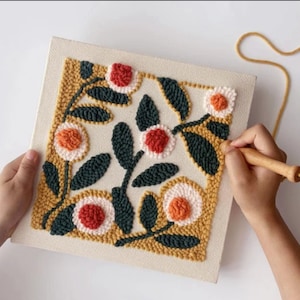 Puede incluir: Una pieza de arte floral con aguja mágica en progreso. El diseño presenta flores con centros rojos y naranjas, rodeadas de pétalos blancos y amarillos, y hojas de color verde oscuro. La pieza está sobre un lienzo cuadrado, sostenido por manos, con una herramienta de aguja mágica de madera.