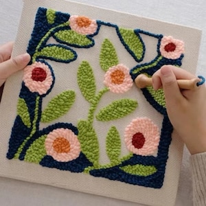 Op de afbeelding: Een close-up van een hand die een houten gereedschap gebruikt om een ​​bloemmotief op een canvas te maken met behulp van de punch needle-techniek. Het ontwerp is gemaakt met roze, groen, blauw en oranje garen.