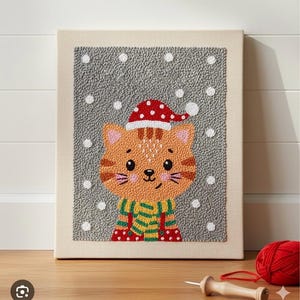 Puede incluir: Una obra de arte enmarcada y cosida a mano que presenta un gato de dibujos animados con un gorro de Papá Noel y una bufanda a rayas. El gato es naranja con rayas marrones, sobre un fondo gris con acentos de copos de nieve blancos.