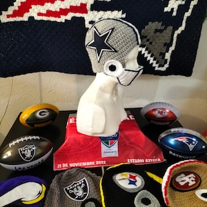 Może przedstawiać: Kolekcja dzianych czapek z logo drużyn futbolowych, w tym Dallas Cowboys, Las Vegas Raiders, Pittsburgh Steelers, San Francisco 49ers i Seattle Seahawks. Czapki są wyeksponowane na czerwono-białym ręczniku z napisem "21 de Noviembre 2022" i "Estadio Azteca".