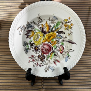 May include: A white ceramic bowl with a floral design. The bowl features a variety of flowers in shades of yellow, pink, red, and blue. The bowl has a scalloped edge and a rope-like design around the rim.