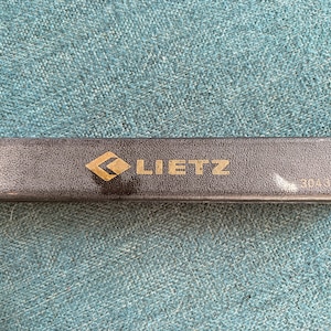 May include: A rectangular, dark gray case with the gold-colored word "LIETZ" and a gold diamond-shaped logo. The case has the number "3043-06" printed on it. The case is resting on a textured, teal surface.