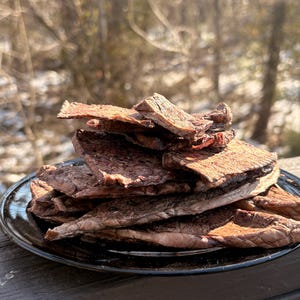 May include: A pile of dark brown beef jerky sits on a black plate. The jerky pieces are stacked, showing a textured surface. The background is blurred, with a glimpse of a snowy landscape and trees.