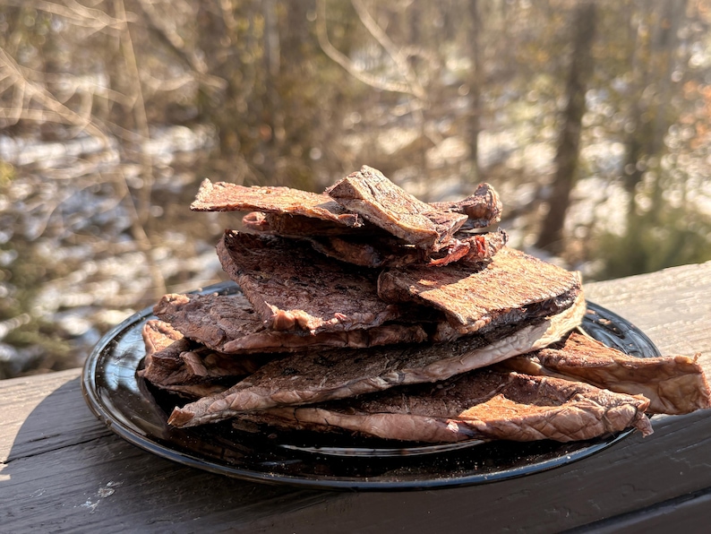 May include: A pile of dark brown beef jerky sits on a black plate. The jerky pieces are stacked, showing a textured surface. The background is blurred, with a glimpse of a snowy landscape and trees.