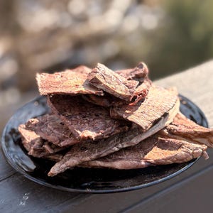 May include: A pile of dried, reddish-brown meat jerky sits on a black plate. The jerky pieces have a textured surface and are arranged in a slightly overlapping manner. The plate is on a wooden surface, with a blurred background.