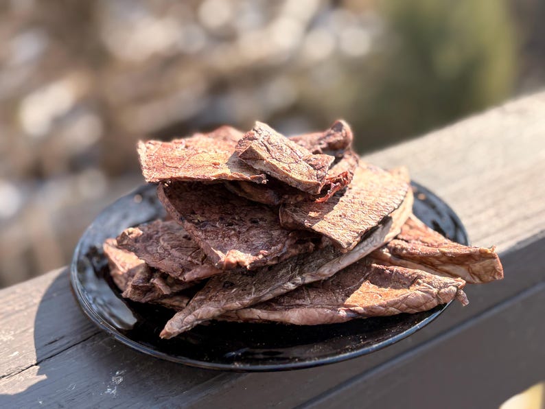 May include: A pile of dried, reddish-brown meat jerky sits on a black plate. The jerky pieces have a textured surface and are arranged in a slightly overlapping manner. The plate is on a wooden surface, with a blurred background.