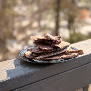 May include: A stack of dark brown, dried food slices on a black plate, resting on a wooden railing. The food appears to be bark-like in texture. The background is blurred, with hints of trees and sunlight.