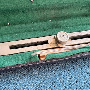 May include: A vintage metal and wood drafting tool, likely for drawing circles, is displayed open in a green felt-lined case. The tool has a sliding mechanism and measurement markings. The case is black with a blue textured background.
