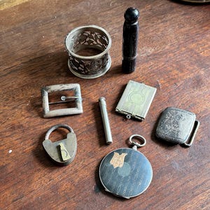May include: A collection of vintage silver and metal trinkets, including a lock, a belt buckle, a small ornate container, a pocket mirror, a small silver container, and a black and silver pocket watch.