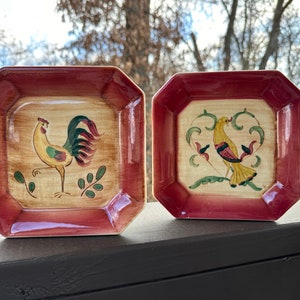 May include: Two small, square, red-rimmed ceramic plates with painted designs. One plate features a rooster with a yellow body, red comb, and green tail feathers. The other plate features a yellow bird with red accents and green leaves.