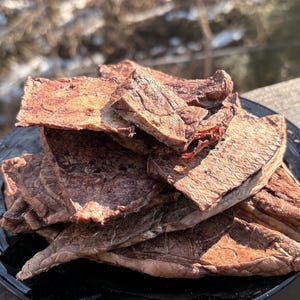 May include: A pile of dried meat jerky on a black plate. The jerky is a reddish-brown color and appears to be thinly sliced. The plate is sitting on a wooden surface, with a blurred outdoor background.
