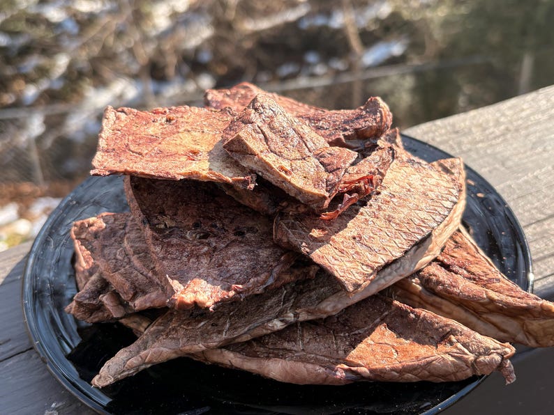 May include: A pile of dried meat jerky on a black plate. The jerky is a reddish-brown color and appears to be thinly sliced. The plate is sitting on a wooden surface, with a blurred outdoor background.