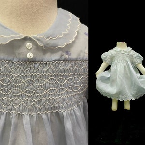 May include: A light blue smocked dress with a white scalloped collar and embroidered flowers. The dress has short puffed sleeves and a gathered skirt.