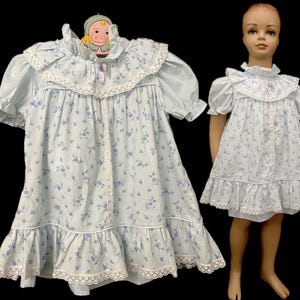 May include: Two white dresses with blue floral patterns and lace trim. The dress on the left has short sleeves and a ruffled hem. The dress on the right has short sleeves and a single ruffle at the hem.