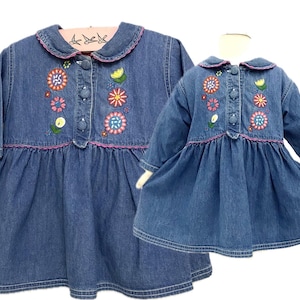 May include: Two denim dresses, one adult and one child-sized, are displayed. Both dresses feature long sleeves, a Peter Pan collar, and a button-down front. The dresses are adorned with colorful floral embroidery and pink trim.