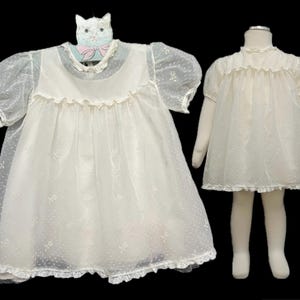 May include: Two white baby dresses. One dress is hanging on a cat-shaped hanger. The dresses have a ruffled neckline and hem. The dresses are made of a sheer fabric with a subtle floral pattern.