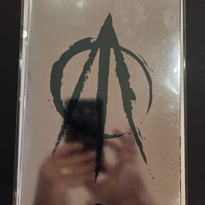 May include: A comic book with a reflective silver metallic cover featuring a large, black, brush-stroke style circular logo in the center. The bottom of the cover displays a small, circular black and white DC logo. The comic is in a clear sleeve.