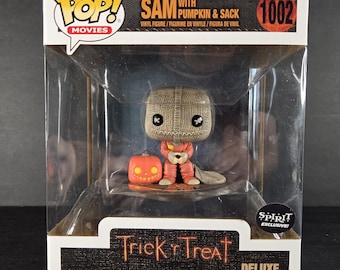 Può includere: Una figura Funko Pop! Movies Deluxe di Sam da Trick 'r Treat, con una zucca e un sacco. La figura è in una scatola espositiva di plastica trasparente con la scritta "Trick 'r Treat" e "DELUXE". La figura indossa un abito arancione.