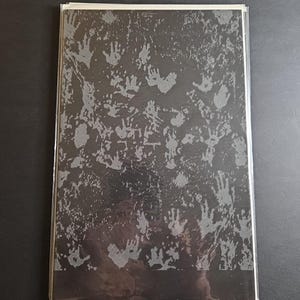 May include: A black and gray graphic print featuring numerous handprints. The design is on a rectangular object, possibly a book or print, encased in a clear plastic sleeve. The handprints are in a repeating pattern.