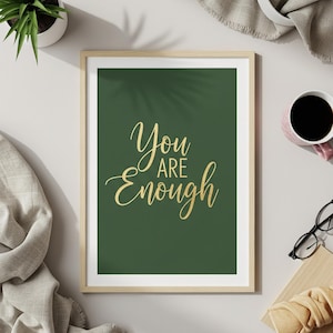 May include: A framed print with the words "You are Enough" in gold script on a dark green background. The print is in a light wood frame. A small potted plant, a cup of coffee, and a pair of glasses are also visible.
