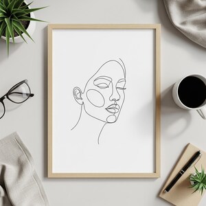 Könnte beinhalten: Ein gerahmter minimalistischer Kunstdruck mit einer Linienzeichnung eines Frauengesichts. Das Kunstwerk ist eine durchgehende Linienzeichnung in Schwarz auf weißem Hintergrund, eingebettet in einen hellen Holzrahmen. Das Bild befindet sich auf einer weißen Oberfläche mit einer Kaffeetasse, einer Brille und einer kleinen Pflanze.